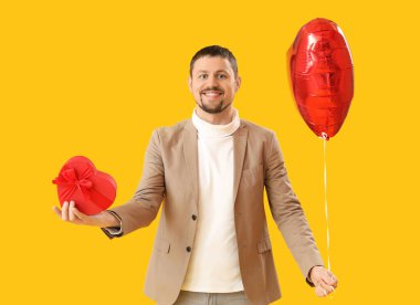 Handsome man with heart-shaped balloon and gift on yellow background. Valentine's Day celebration
