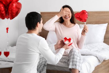 Young man surprising his girlfriend with gift in bedroom on Valentine's Day