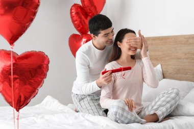 Young man surprising his wife with gift in bedroom on Valentine's Day