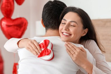 Young woman with gift hugging her boyfriend in bedroom on Valentine's Day, closeup