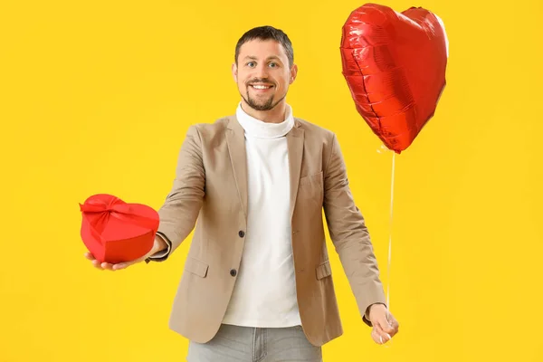Handsome man with heart-shaped balloon and gift on yellow background. Valentine\'s Day celebration