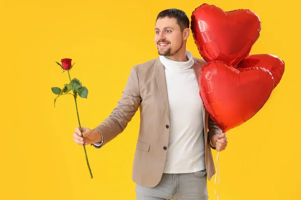 Handsome man with rose and heart-shaped balloons on yellow background. Valentine\'s Day celebration