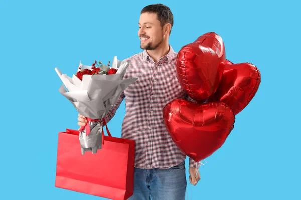 Handsome man with heart-shaped balloons, shopping bag and flowers on blue background. Valentine\'s Day celebration