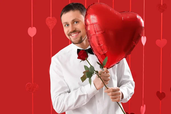 Handsome man with heart-shaped balloon and rose on red background. Valentine\'s Day celebration