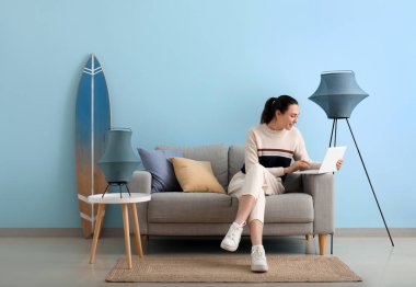 Mavi duvarın yanındaki gri koltukta dizüstü bilgisayar kullanan genç bir kadın.