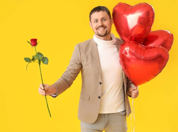 Handsome man with rose and heart-shaped balloons on yellow background. Valentine\'s Day celebration