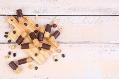 Cutting board with heap of delicious wafer rolls, coffee beans and nuts on light wooden background