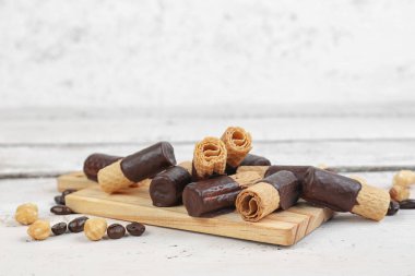 Cutting board with heap of delicious wafer rolls, coffee beans and nuts on light wooden background, closeup