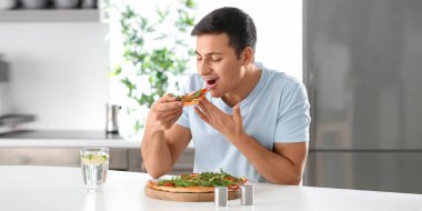 Young man eating tasty pizza in kitchen
