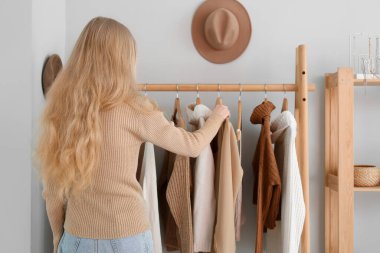 Blonde woman and rack with stylish clothes near light wall