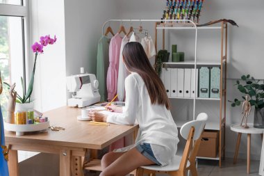 Female tailor with cup of coffee drawing sketch at table in atelier
