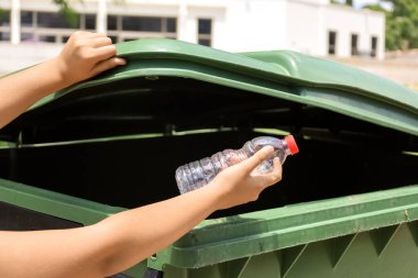 Child throwing plastic bottle into garbage container outdoors, closeup