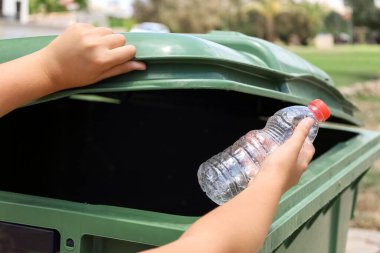 Child throwing plastic bottle into garbage container outdoors, closeup