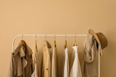 Rack with stylish clothes and hat near beige wall
