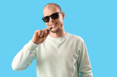 Young man in sunglasses with disposable electronic cigarette on blue background