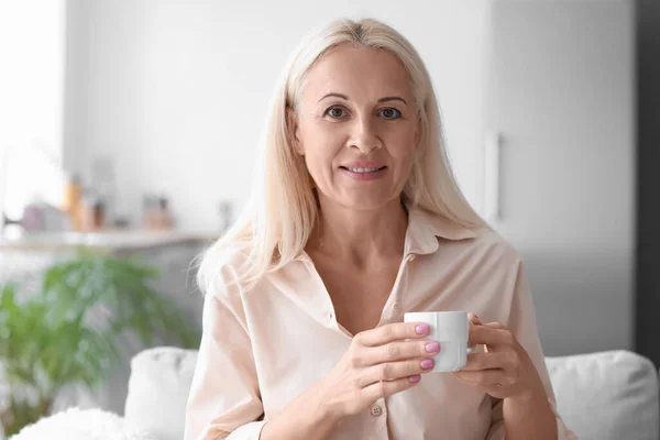 Mature woman with cup of hot beverage sitting on sofa at home, closeup