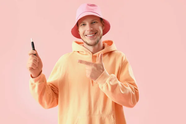 Young man in bucket hat pointing at disposable electronic cigarette on pink background
