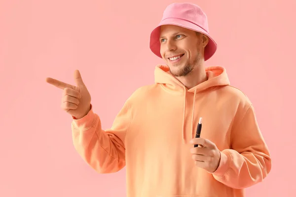 Young man in bucket hat with disposable electronic cigarette pointing at something on pink background