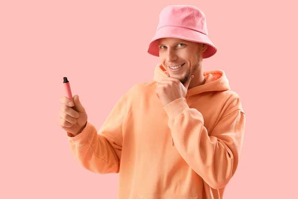 Thoughtful young man in bucket hat with disposable electronic cigarette on pink background