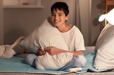 Little boy with pillow sitting on electric heating pad in bedroom at night