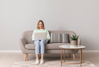 Gri duvarın yanındaki yumuşak koltukta dizüstü bilgisayar kullanan genç bir kadın.