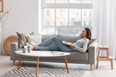 Genç bir kadın oturma odasındaki gri koltukta dizüstü bilgisayar kullanıyor.