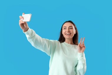 Young woman with mobile phone taking selfie on blue background