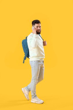 Young bearded man with backpack talking by mobile phone on yellow background