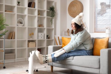 Frozen young woman in down jacket warming near radiator at home
