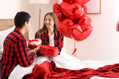 Young couple greeting each other with gifts in bedroom on Valentine's Day