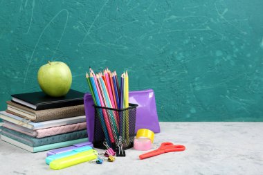 Holder with stationery, notebooks, pencil case and apple on table near blackboard