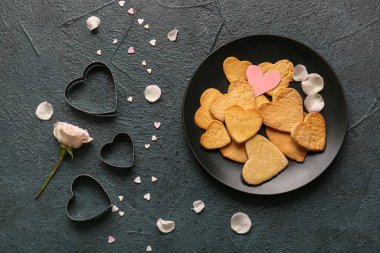 Composition with plate of tasty heart shaped cookies, cutters and rose petals on dark color background. Valentines Day celebration