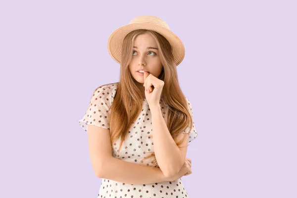 Young woman in hat biting nails on lilac background