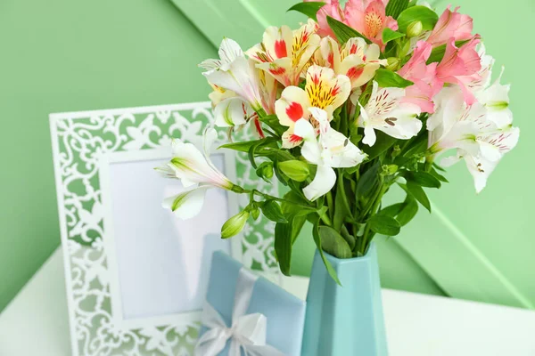 Vase with beautiful alstroemeria flowers, gift and picture frame on table near color wall, closeup. Mother\'s day celebration