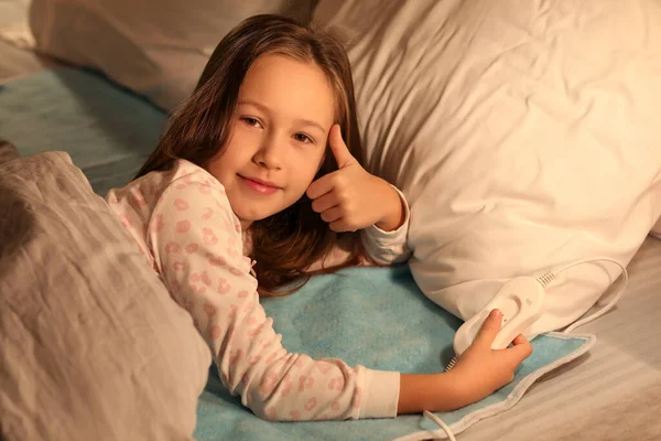 Little girl with electric heating pad showing thumb-up in bedroom at night