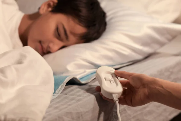 Mother turning on electric heating pad for her little boy in bedroom at night, closeup
