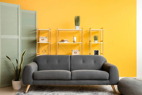 Interior of living room with folding screen, cozy sofa and shelving units near yellow wall