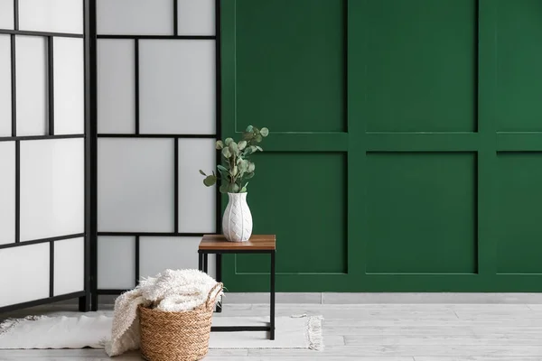 Folding screen, eucalyptus in vase and plaid in basket near green wall