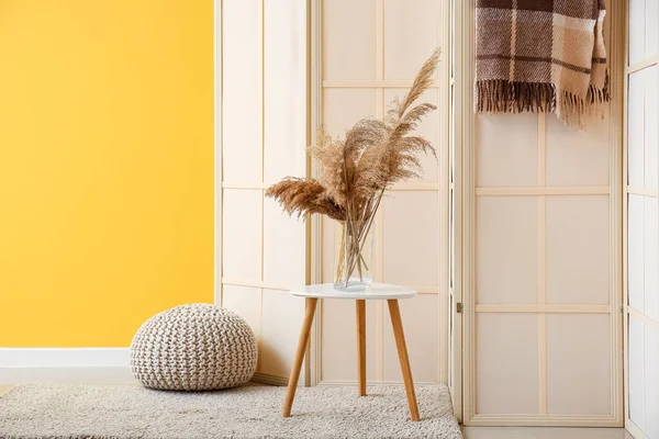 Beige folding screen, pampas grass in vase and pouf near yellow wall