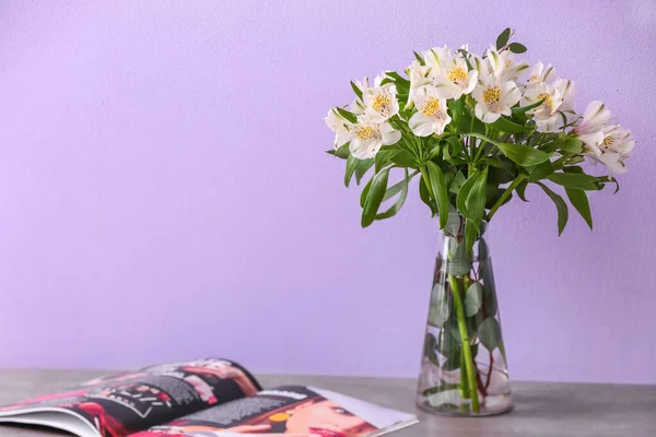 Vase with bouquet of alstroemeria flowers and magazine on table near purple wall