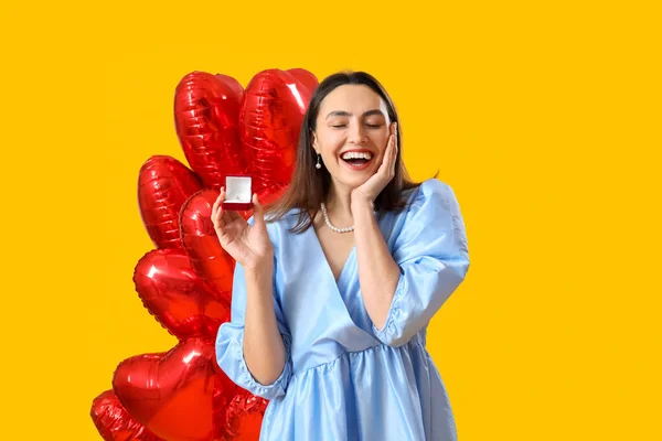 Happy young woman with engagement ring and balloons on yellow background. Valentine\'s Day celebration