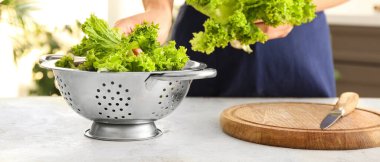 Woman putting lettuce leaves into colander on table in kitchen