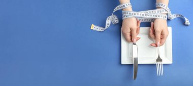 Female hands tied with measuring tape, empty plate and cutlery on blue background with space for text. Concept of diet