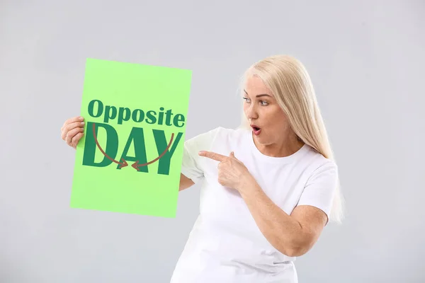 Shocked mature woman holding paper sheet with text NATIONAL OPPOSITE DAY on light background