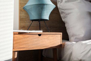 Wooden bedside table with lamp and magazine in bedroom, closeup