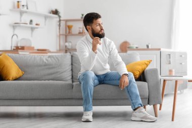 Thoughtful young bearded man sitting on sofa in kitchen