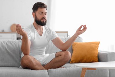 Handsome young man meditating on sofa at home