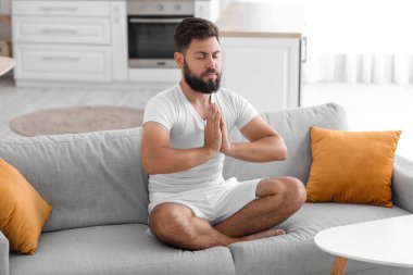 Handsome young man meditating on sofa at home