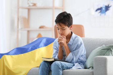 Little boy with Holy Bible and flag of Ukraine praying at home