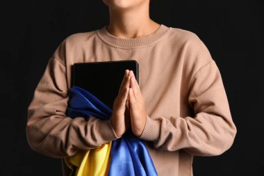 Little boy with Holy Bible and flag of Ukraine praying on black background, closeup